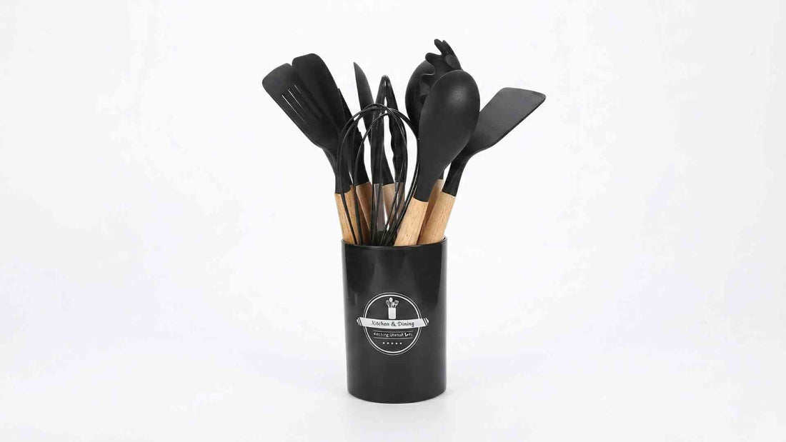 A set of black silicone and wooden cooking utensils, including spatulas, a whisk, and a pasta server, standing upright in a black cylindrical holder labeled "Kitchen & Dining Cooking Utensil Sets."