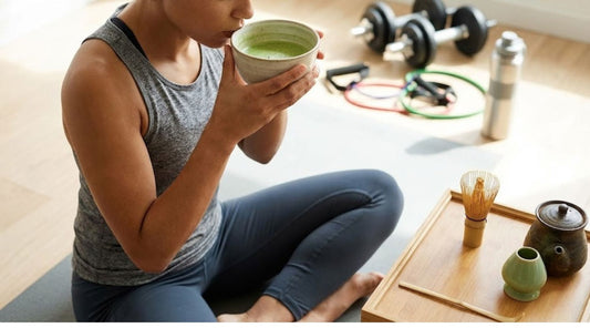Woman in athletic gear drinks matcha on a yoga mat near dumbbells and a traditional tea set.