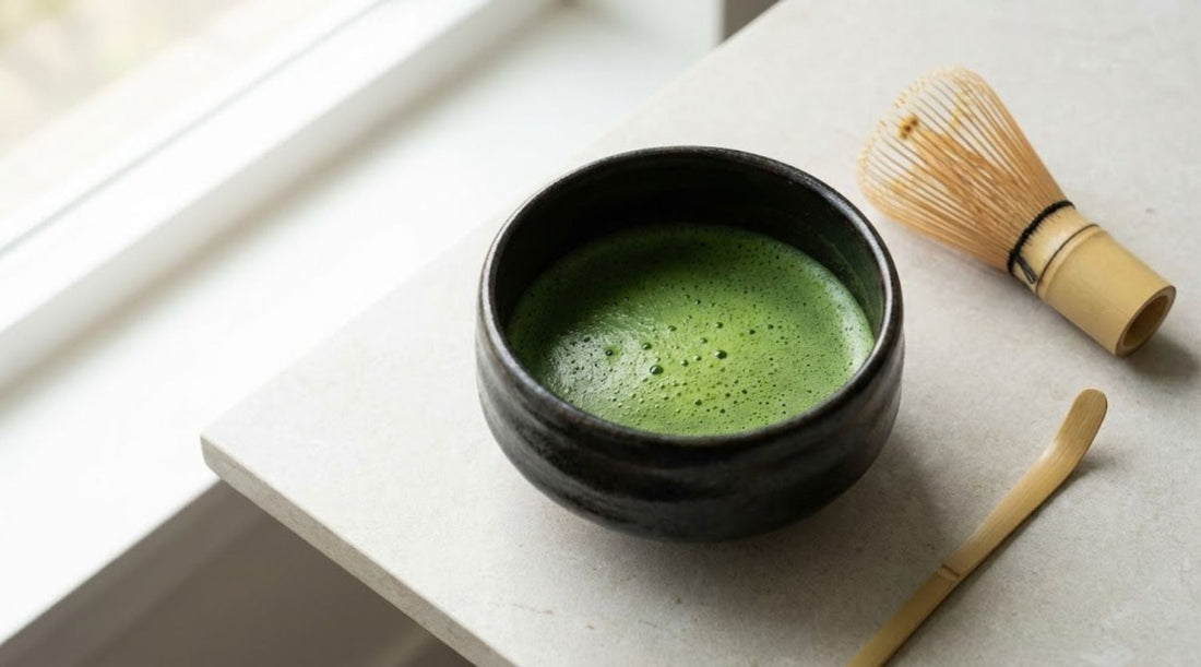 A dark ceramic bowl of frothy matcha tea sits on a light stone countertop next to a bamboo whisk and scoop in bright natural light.