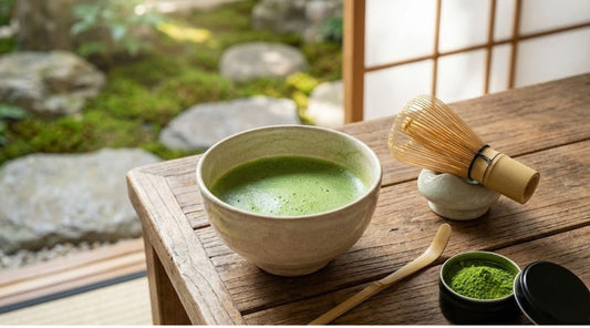 A bowl of frothed matcha with a bamboo whisk and scoop on a table by a Zen garden.