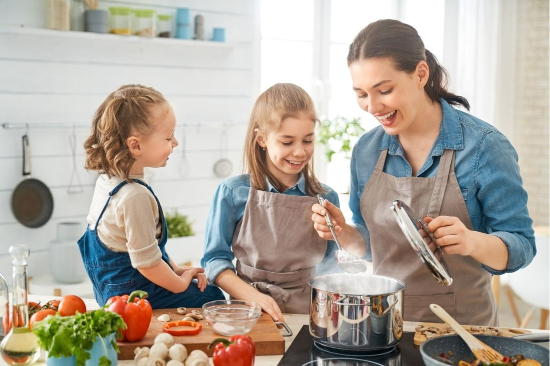 A woman and two young girls smiling and cooking together in a bright, modern kitchen. The woman is lifting the lid of a steaming pot on the stove, while a variety of fresh vegetables like bell peppers, tomatoes, and mushrooms are laid out on the counter.