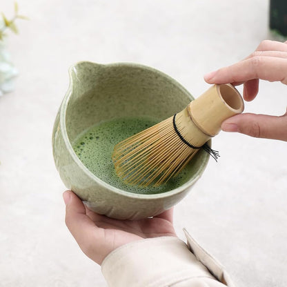 Person holding a green ceramic bowl with matcha and a bamboo whisk on a light background